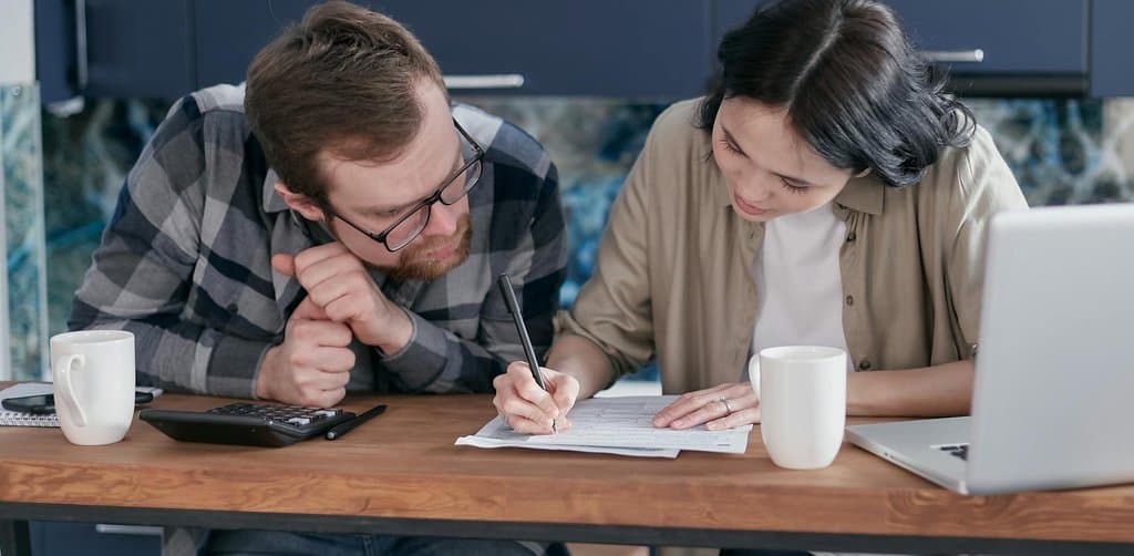 A bearded man in a checkered shirt and a woman in a beige cardigan sitting close together at a kitchen table reviewing a paper document with a laptop and calculator in front of them, the kind of family-level financial conversation a borrower should have before signing an SBA loan personal guarantee