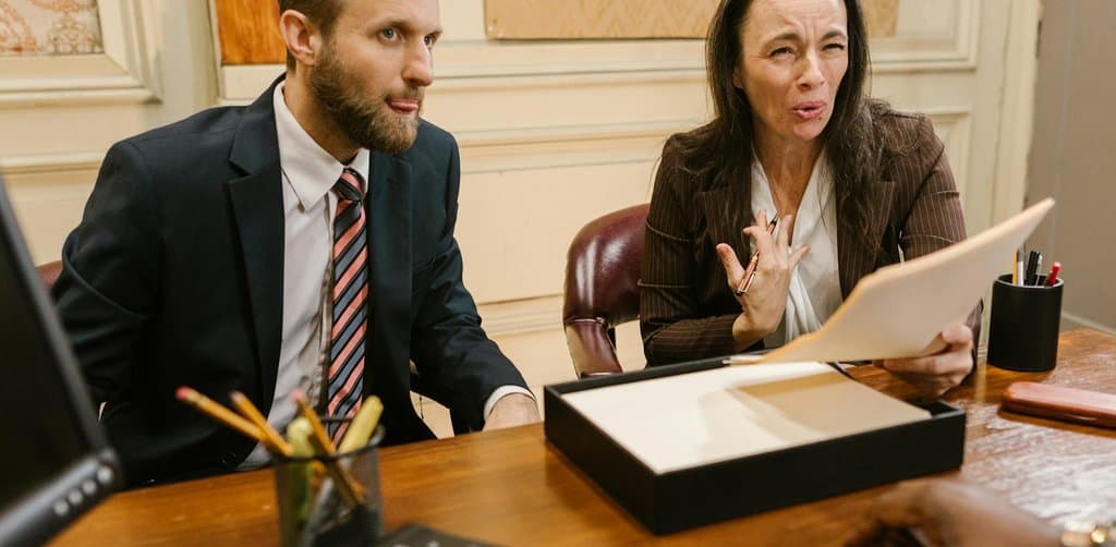 A man in a navy suit and striped tie alongside a woman in a brown blazer holding a folder of documents, both seated at a polished wooden desk having a serious conversation about SBA loan personal guarantee terms