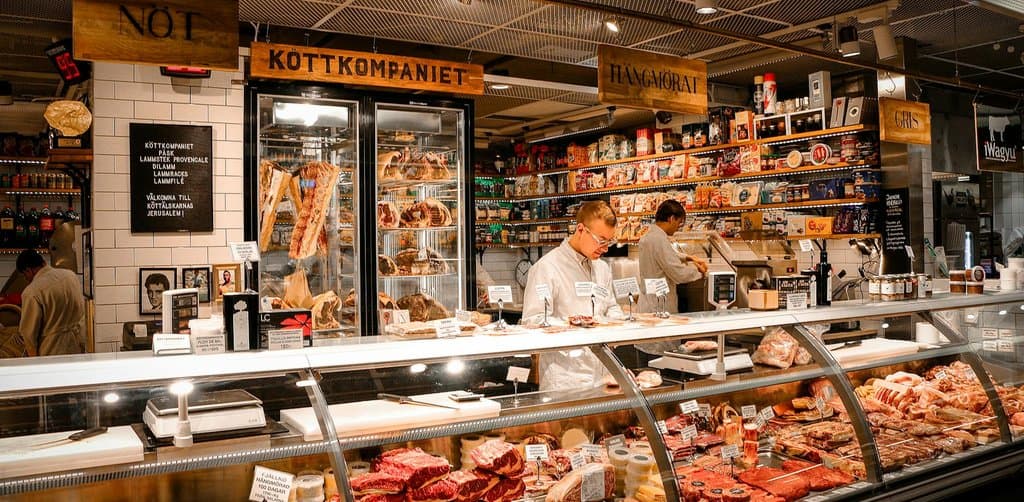 A bright butcher and deli counter labeled KOTTKOMPANIET with a butcher in white standing behind glass display cases full of cured meats and prepared foods inside an indoor market, the kind of small business where SBA seller financing requirements frequently appear in the closing structure