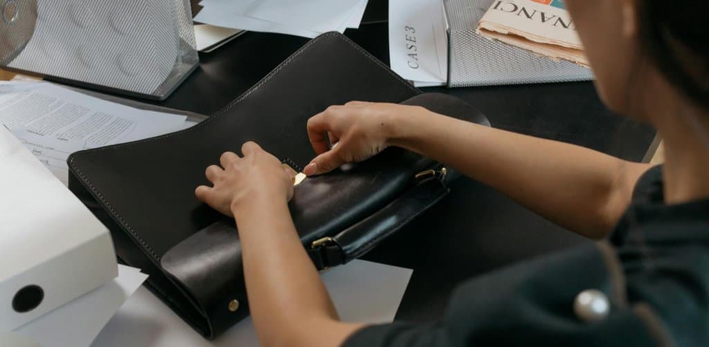 A woman in a dark top working at a cluttered desk packing a black leather briefcase amid loose paperwork, an open document tray labeled CASE 3, and a mesh inbox stacked with newspapers, the closing-table preparation for an SBA-financed acquisition with seller financing in the capital stack