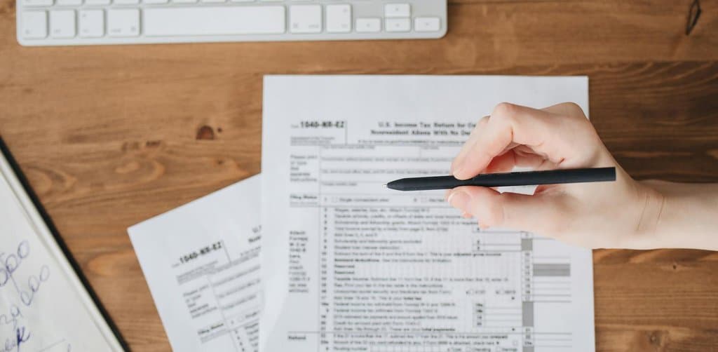 Top-down view of a hand holding a black pen over a printed US 1040-NR-EZ income tax return form on a wooden desk with a white keyboard partially visible above and handwritten financial notes on a clipboard to the side, the federal filing where seller financing tax implications get formally reported