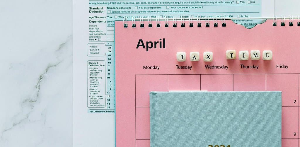 A pink April-themed planner labeled APRIL TAX TIME with small wooden tiles spelling out TAX TIME laid on a partially visible 1040 individual income tax form against a marble background, the tax season urgency that defines seller financing tax implications planning