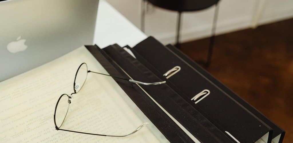 Three thick black ring binders bound with metal clip spines stacked at the right edge of a white desk next to a silver laptop with a pair of round wire-rimmed reading glasses resting on top of an open page of typed text under warm soft lighting, the documentation package every buyer of a note secured by business assets demands before pricing the paper