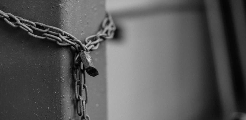 A close-up black and white photograph of a thick metal chain wrapped tightly around a painted steel post and secured by a heavy rusted iron padlock with the chain end hanging down against the textured metal surface in soft natural light, the perfected security interest that backs every note secured by business assets