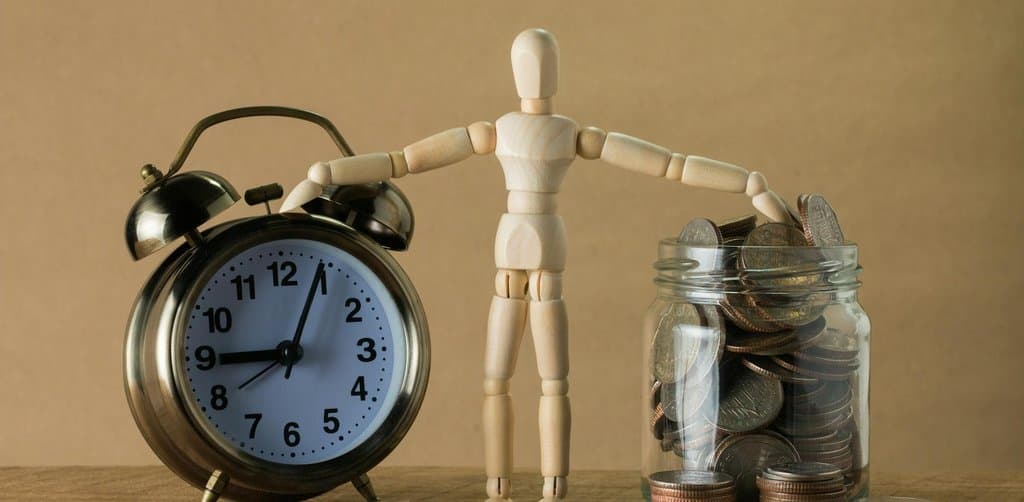 A wooden artist's mannequin figure standing between a brass twin-bell alarm clock on the left and a glass jar overflowing with silver and copper coins on the right with loose coins scattered on a wooden surface, the time-value-of-money math that drives every business note pricing conversation in the secondary market