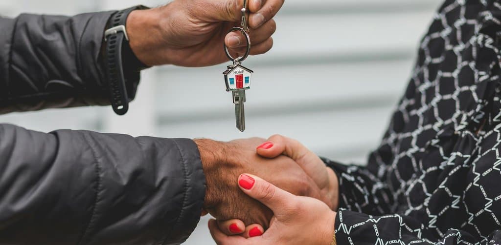 Two hands meeting in handshake position while one hand passes a metal house key with a small red and blue HOME charm attached to the keyring on the seller's keychain to the buyer's hand against a gray clapboard background, the moment of transfer that closes every seller-financed business note sale