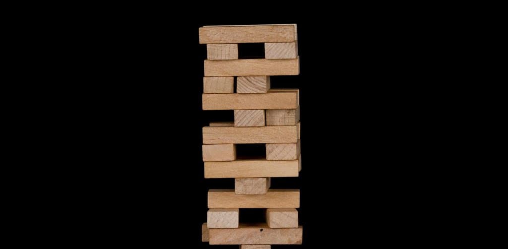 A vertical wooden block tower made from stacked rectangular pieces of pale wood arranged like a Jenga puzzle photographed against a solid black background, the visual metaphor for the layered capital stack that puts subordinated seller notes above buyer equity but below senior bank debt