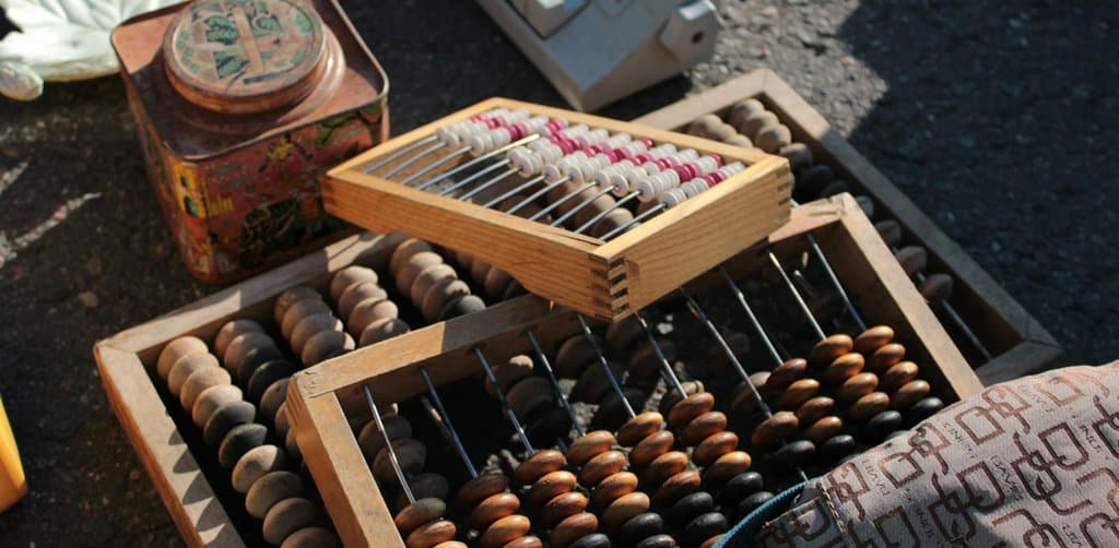 An assortment of vintage Russian wooden abacuses with large round wood beads in chestnut and black sit in shallow open wooden display crates on dark asphalt at an outdoor flea market beside an antique red metal tea tin with floral motif and an old white plastic mechanical calculator, the traditional counting tools that frame the careful comparison work owners need to do across types of business loans