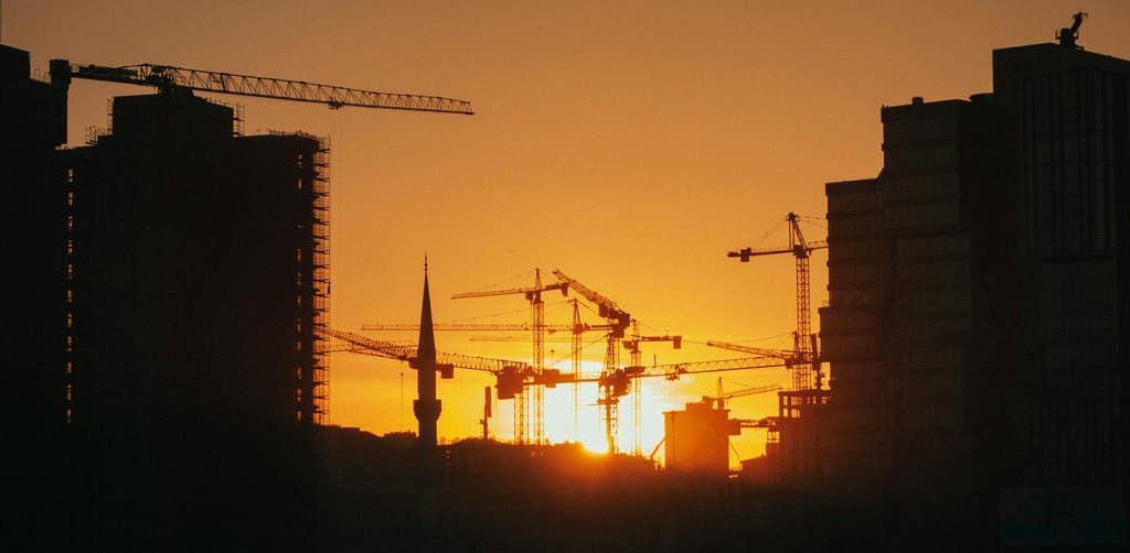 A line of tall construction tower cranes stand silhouetted against a deep amber-orange sunset sky in a busy city skyline with two unfinished concrete tower buildings flanking the frame and a single minaret rising between the cranes near the setting sun, the active mid-build commercial real estate moment that draws on long-term SBA 504 and commercial mortgage types of business loans