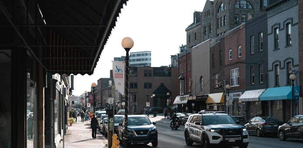 A late-afternoon view of a small-town main street with a row of independent shops, a historic red-brick building in the background, parked cars along the curb, and a police vehicle driving down the road, the kind of small business district that SBA loans fund across the United States